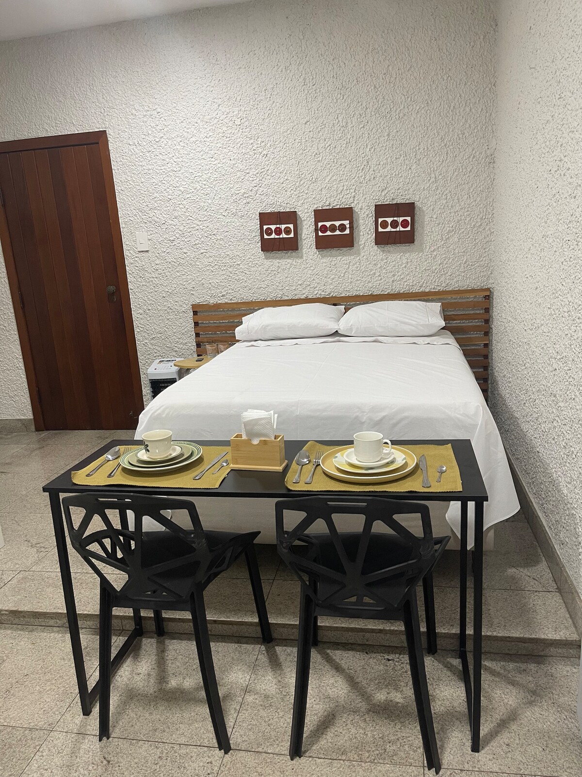 A cozy room features a spacious bed with white linens against a textured wall. A small dining table is set with two place settings, including cups and utensils. A wooden door and framed artwork add warmth to the modern design.