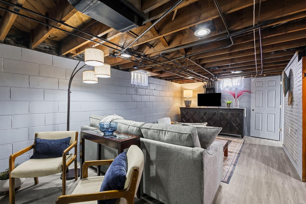 A spacious basement area features comfortable seating arranged for relaxation. Natural light filters in through small windows, illuminating the seating and accent decor. A large television is positioned on a stand against the wall, complemented by a stylish bar area and decorative elements.