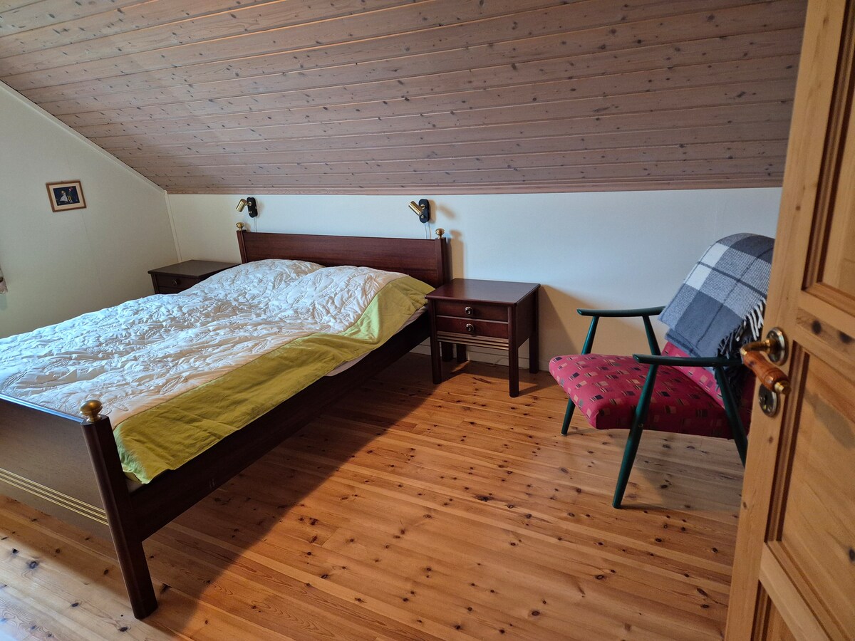 غرفة نوم مواجهة للشرق بسرير مزدوج بعرض 150 سم وألحفة بطول 220 سم. منضدتان ومصباحان.