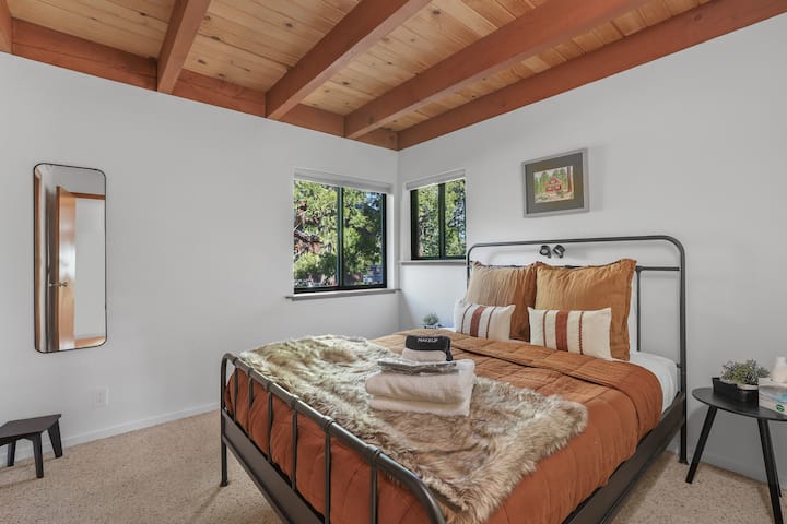 Cozy bedroom with a comfortable queen bed, soft layered linens, and exposed wood-beam ceilings. Large windows bring in natural light and peaceful tree views for a restful night’s sleep.