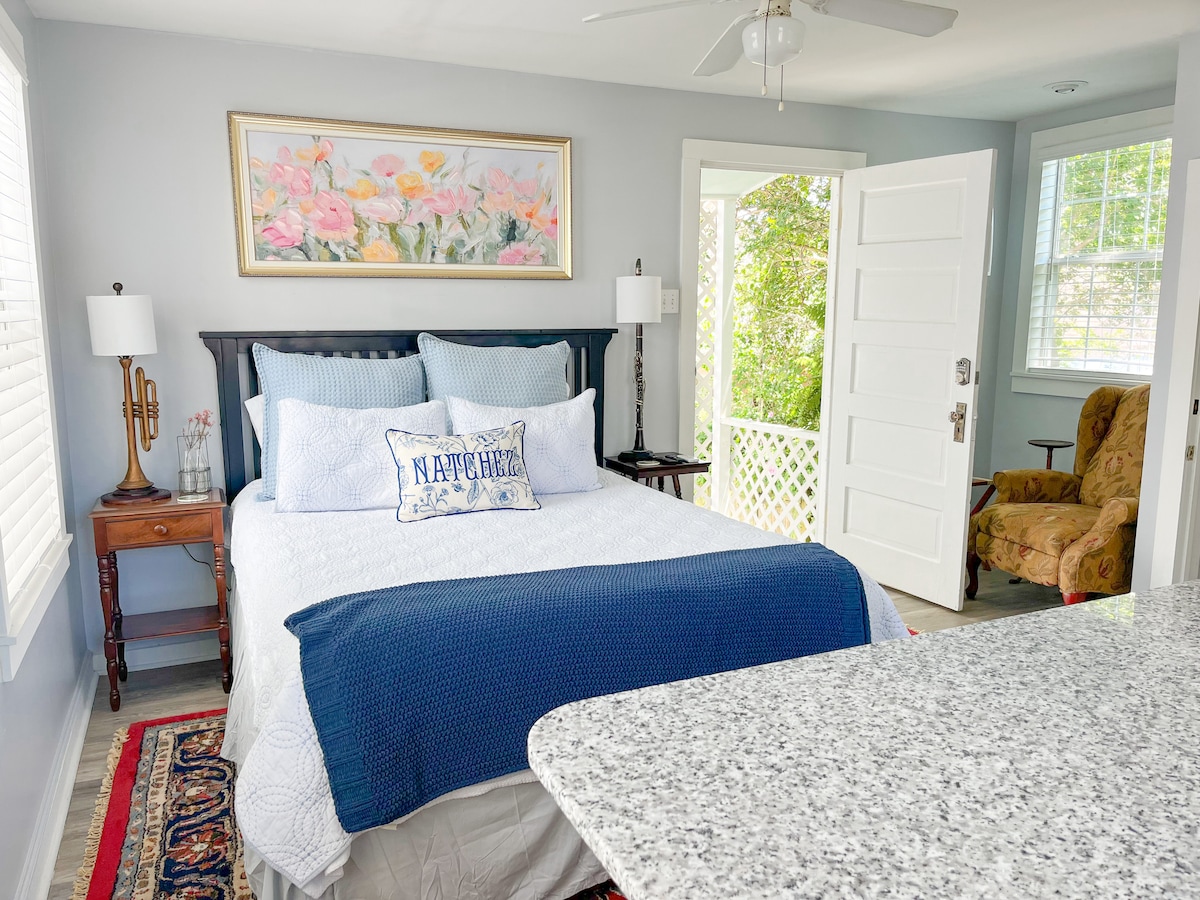 A well-lit bedroom features a spacious bed with white linens and a dark blue throw blanket. Two bedside lamps provide soft illumination beside the bed. A floral painting adorns the wall above, and a cozy chair sits near an open door leading to a small balcony.