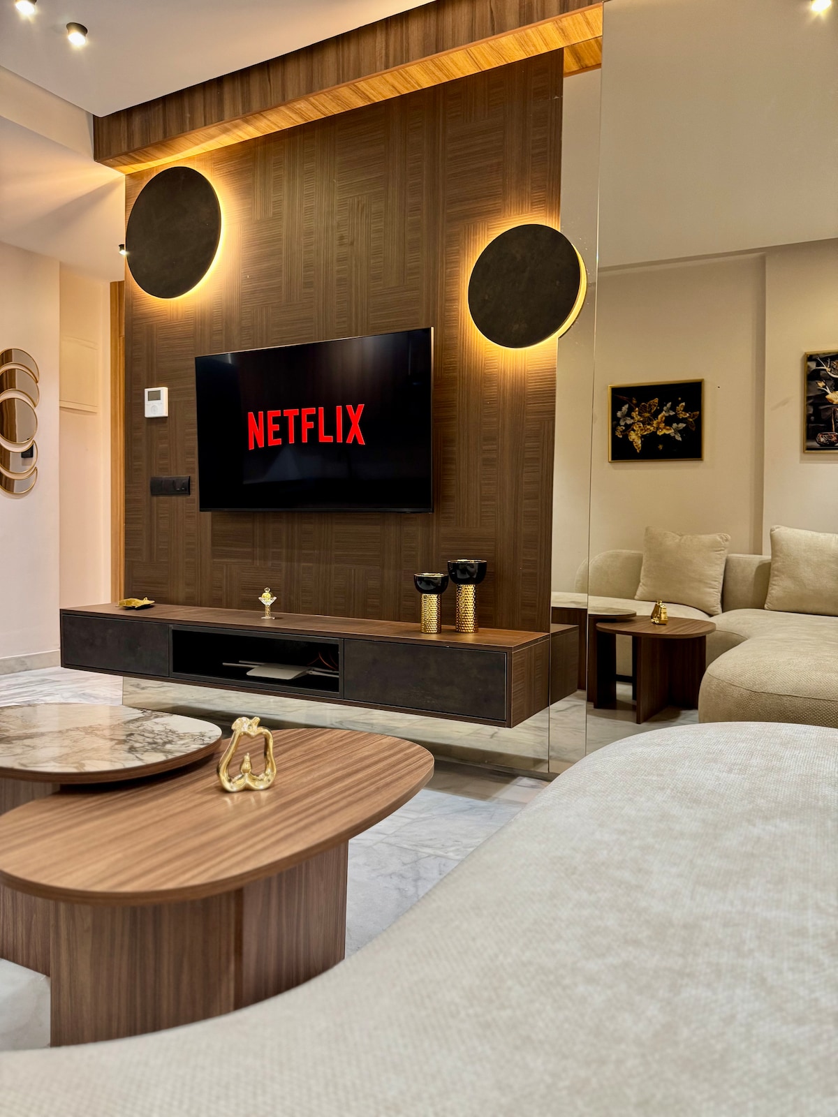 A modern living area features a large wall-mounted television displaying the Netflix logo. Two wooden tables with round and rectangular shapes sit in front of a plush light-colored sofa. Soft lighting complements the warm tones of the wood paneling, enhancing the inviting space.