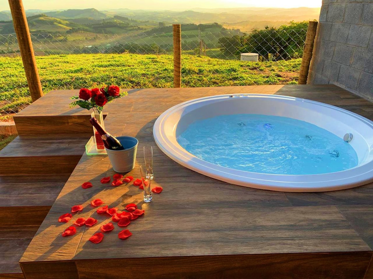 A private jacuzzi is positioned on a wooden deck, surrounded by rose petals. A bouquet of red roses and two flutes are placed nearby, alongside a bottle of champagne. The serene landscape of rolling hills is visible in the background, enhancing the tranquil atmosphere.