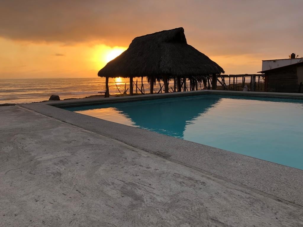 A tranquil pool reflects the warm hues of the setting sun, with a thatched-roof pavilion situated nearby. The smooth concrete surrounds the pool, creating a spacious area for relaxation while the ocean is visible in the background.