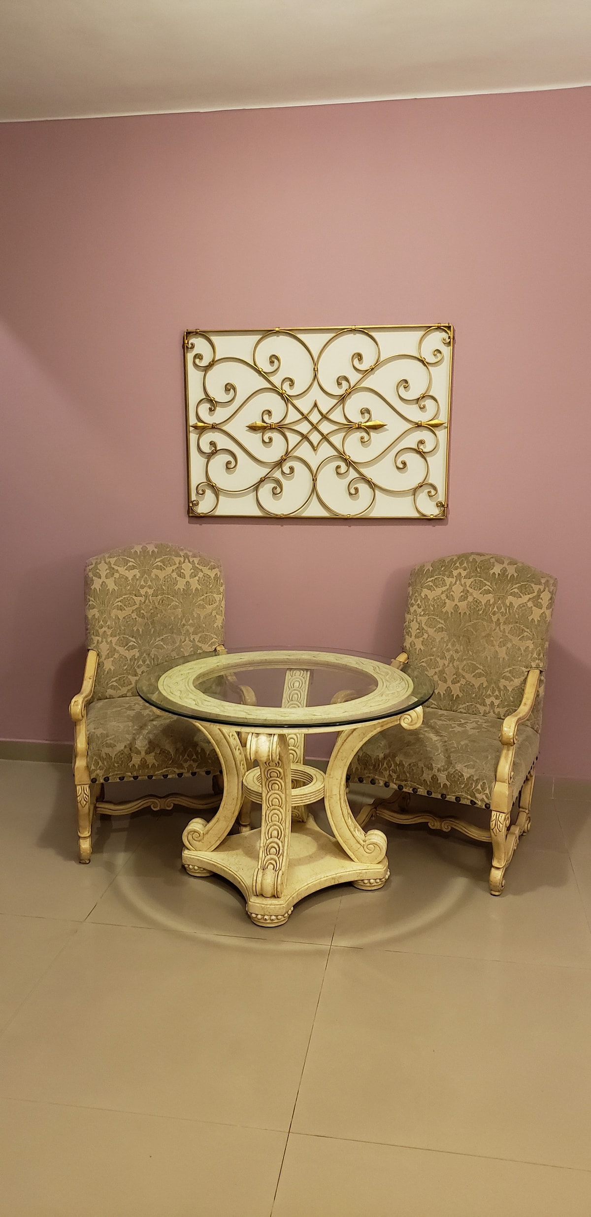 An elegant seating arrangement is presented, featuring two patterned chairs flanking a round glass table. The soft color palette of the wall enhances the charm of the space. A decorative wall piece adds a subtle artistic touch.