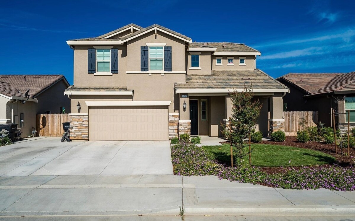 A two-story home is presented, featuring a light beige exterior with dark blue shutters. A spacious lawn is surrounded by colorful landscaping, while a double garage is visible to the left. The driveway offers ample parking space, contributing to the home's welcoming ambiance.