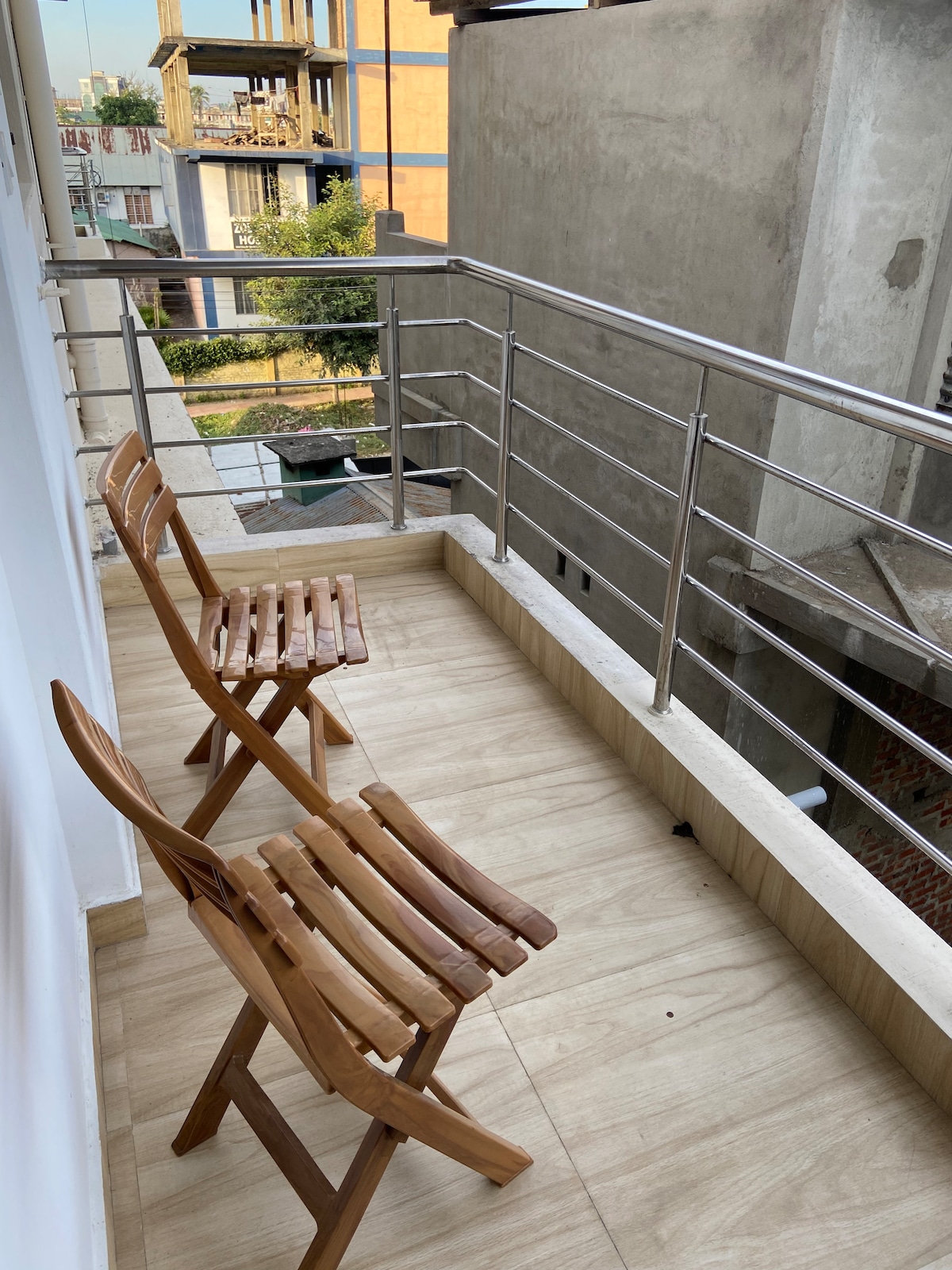 An outdoor balcony is showcased with two wooden folding chairs positioned against a railing. The floor is finished with light-colored tiles, and views of nearby buildings and greenery are visible in the background.