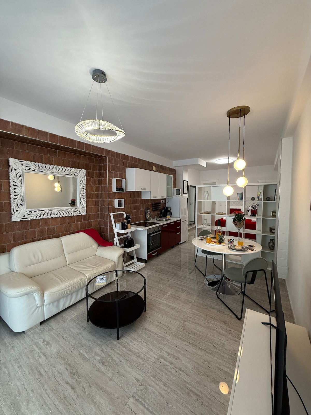 An open-plan living area features a white leather sofa and a round dining table set for four. A stylish mirror hangs above a dark cabinet, with a well-equipped kitchen visible. Soft lighting is provided by modern fixtures, creating a bright and welcoming ambiance.