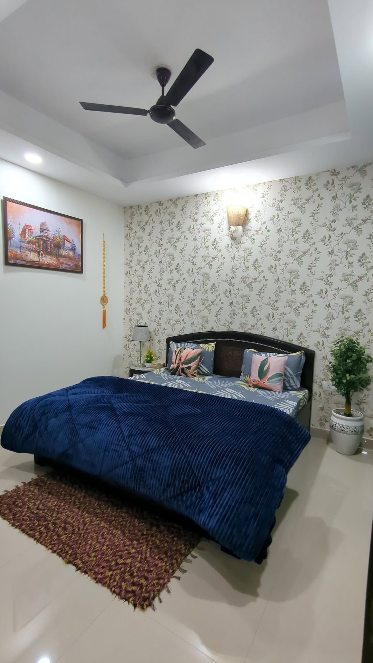 A cozy bedroom features a spacious bed adorned with a soft blue blanket, complemented by decorative cushions. Floral wallpaper adds a touch of elegance, while a ceiling fan and wall light provide illumination. A small planter and a rug enhance the inviting atmosphere.