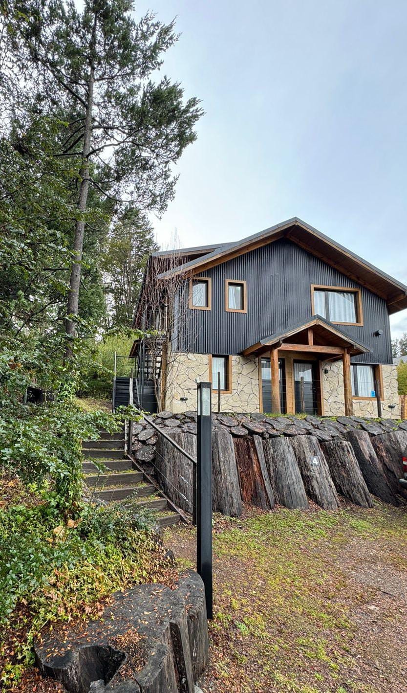 A two-story house with a dark exterior is nestled amidst greenery. A stone pathway leads to the entrance, flanked by natural wood landscaping. Tall trees provide shade, and large windows invite light into the living spaces. The overall setting conveys a serene atmosphere.