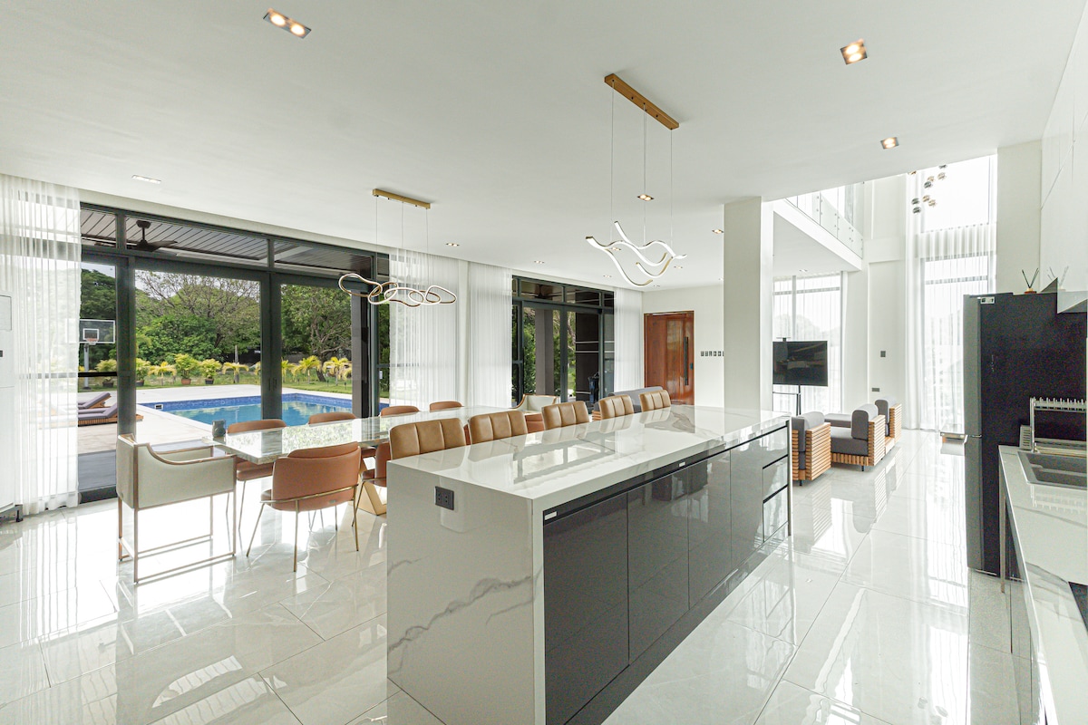An open-concept living and dining area showcases modern furnishings and ample natural light. A large dining table is positioned beside a sleek kitchen island, while floor-to-ceiling windows reveal views of the swimming pool and surrounding greenery.
