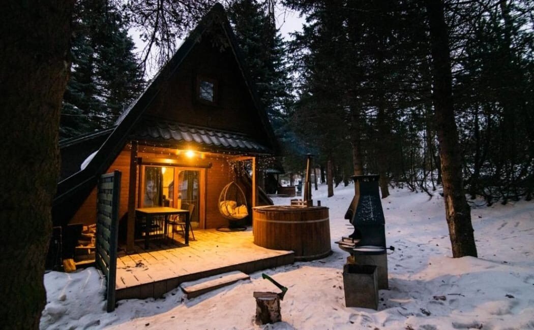 A charming cabin is nestled among snow-covered trees, featuring warm lighting that creates a welcoming ambiance. A wooden hot tub is positioned on the deck, alongside a swing chair and outdoor dining area, allowing for relaxation in a serene setting.