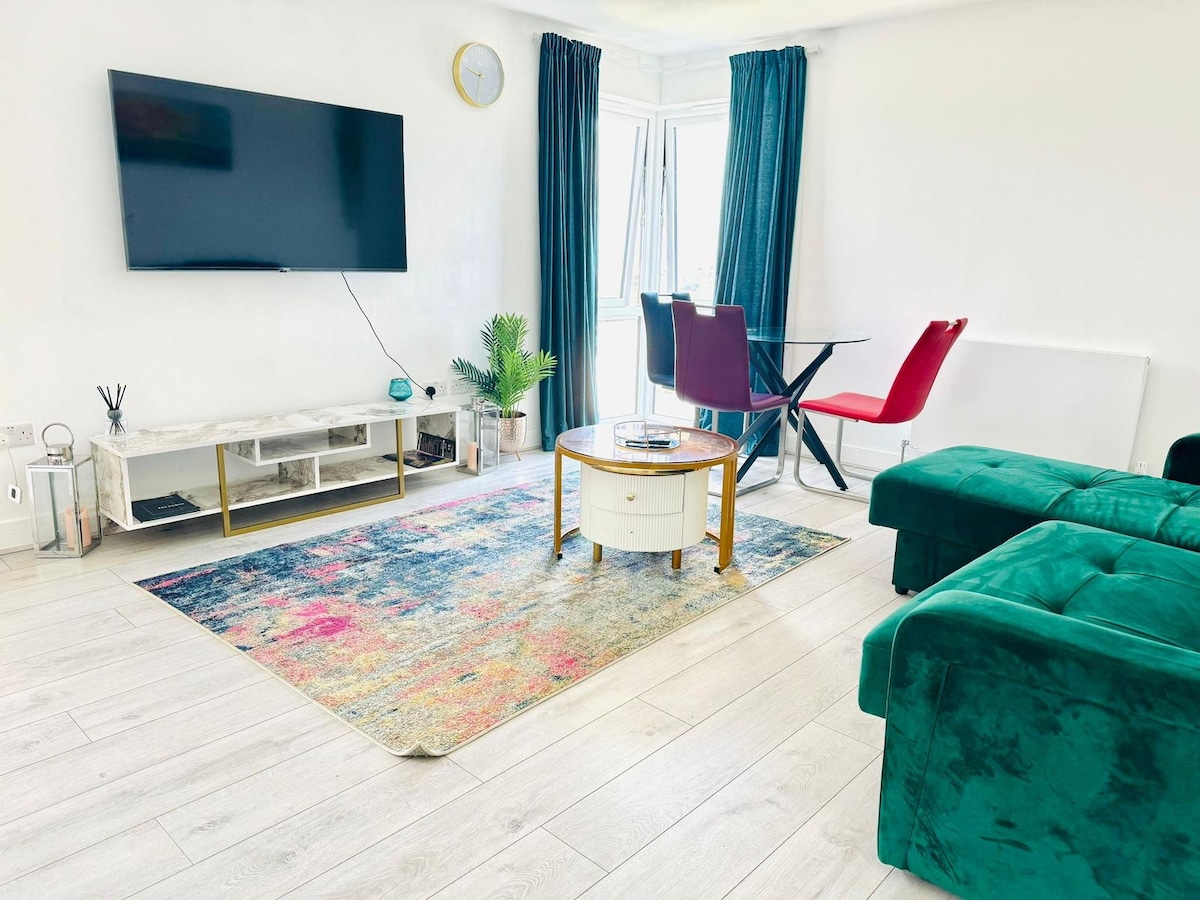 A modern living area features a large flat-screen TV mounted on the wall, complemented by a stylish coffee table centered on a colorful area rug. A dining table with red and black chairs sits nearby, while large windows are framed by dark curtains, allowing natural light to fill the space.