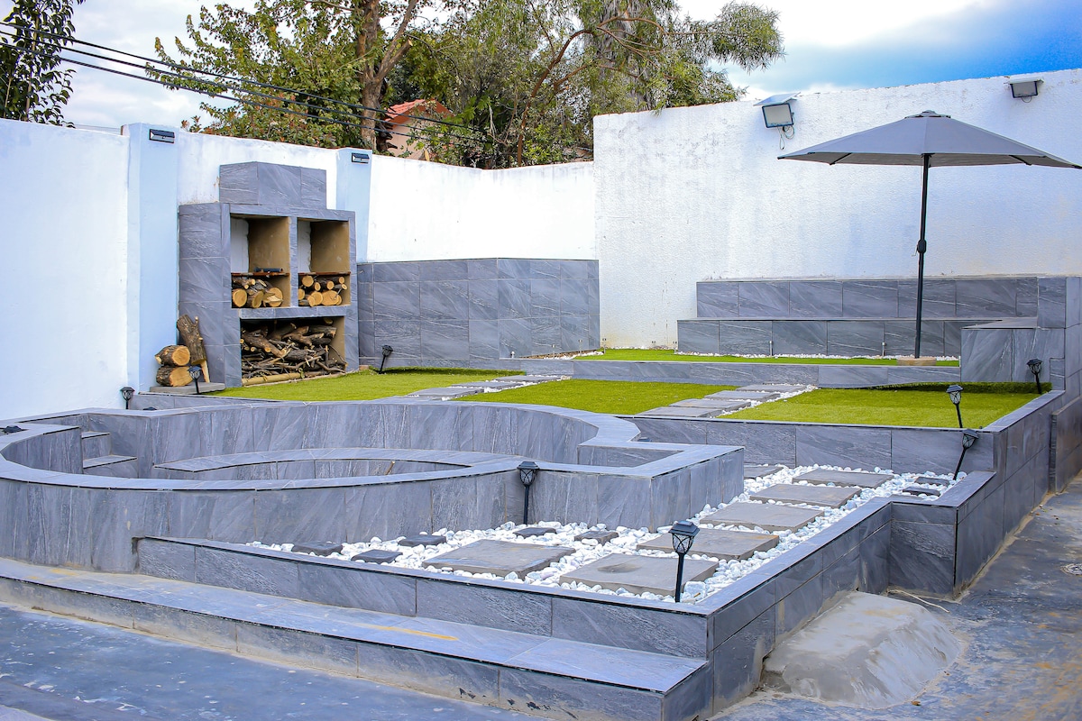 An outdoor area features modern stone landscaping with interspersed grass patches. A shaded seating area is positioned under a large umbrella, while stacked logs are neatly arranged beside a built-in fire pit. Subtle path lighting guides through the space.