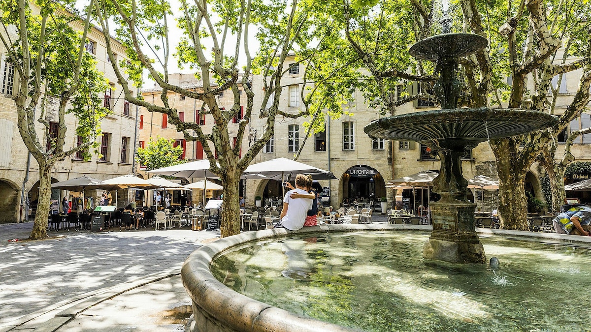 A communal square is centered around a stone fountain, surrounded by trees providing shade. Outdoor seating under umbrellas offers a place for diners to relax, while charming buildings with balconies frame the scene, enhancing the village's tranquil atmosphere.