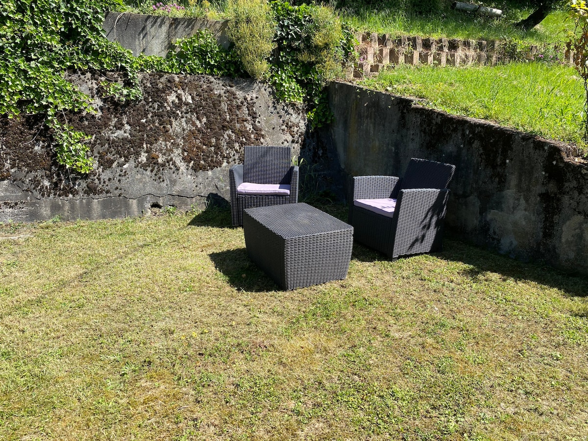 A private garden area is featured, showcasing two woven chairs with cushions and a low table. The grassy space is bordered by a moss-covered stone wall and lush greenery, providing a serene setting for relaxation.