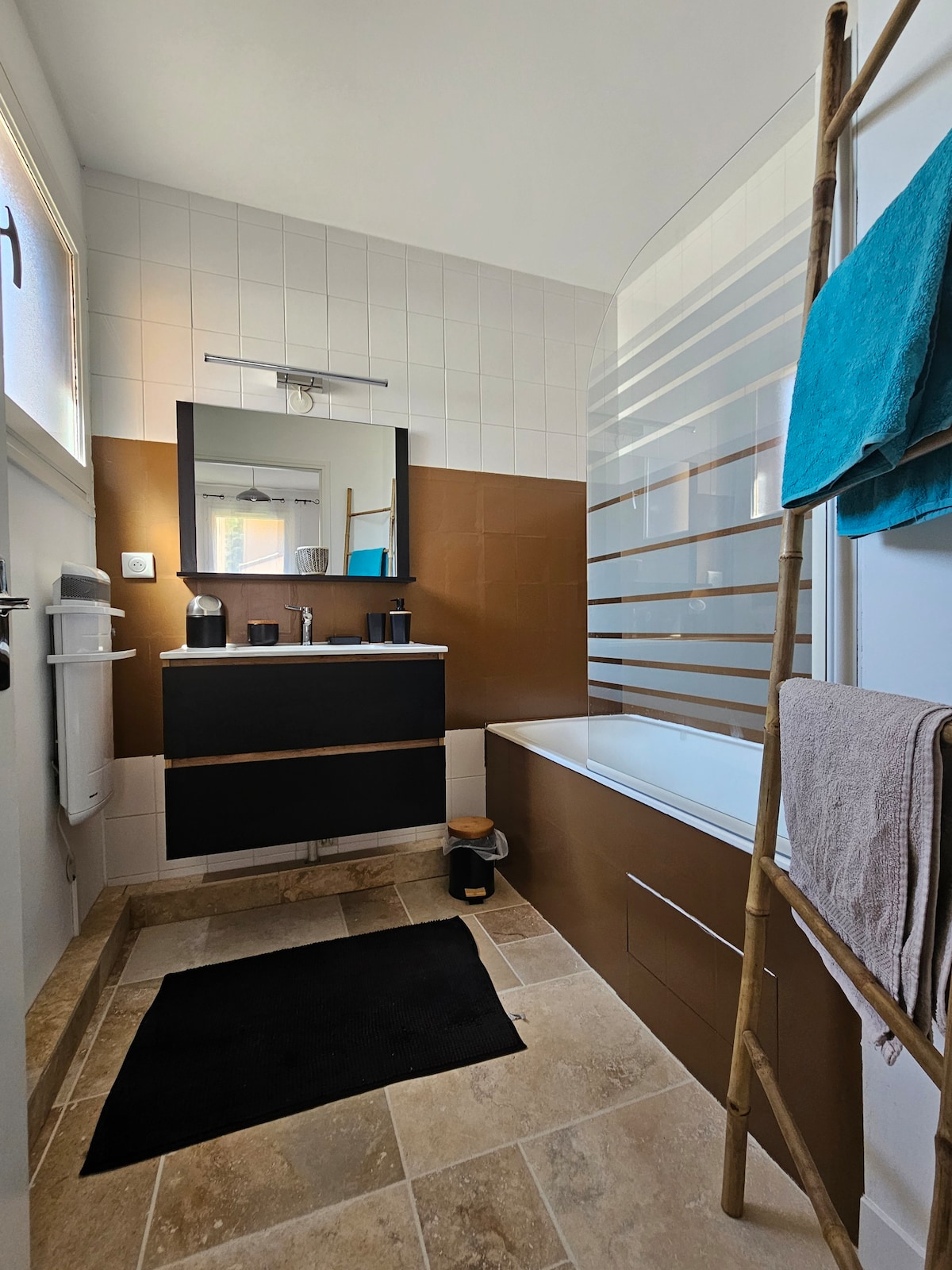 The bathroom features a bathtub alongside a modern vanity with a mirror. A towel rack made of bamboo displays two towels. The floor is covered with neutral tiles, and a dark mat is positioned in front of the sink area, creating a functional and organized space.