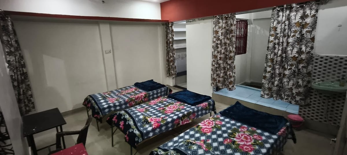 A shared sleeping area contains three spacious beds, each covered with colorful floral blankets. Flowery curtains provide privacy, while a small table can be seen in the corner. A washbasin is visible near the back, contributing to the functionality of the space.