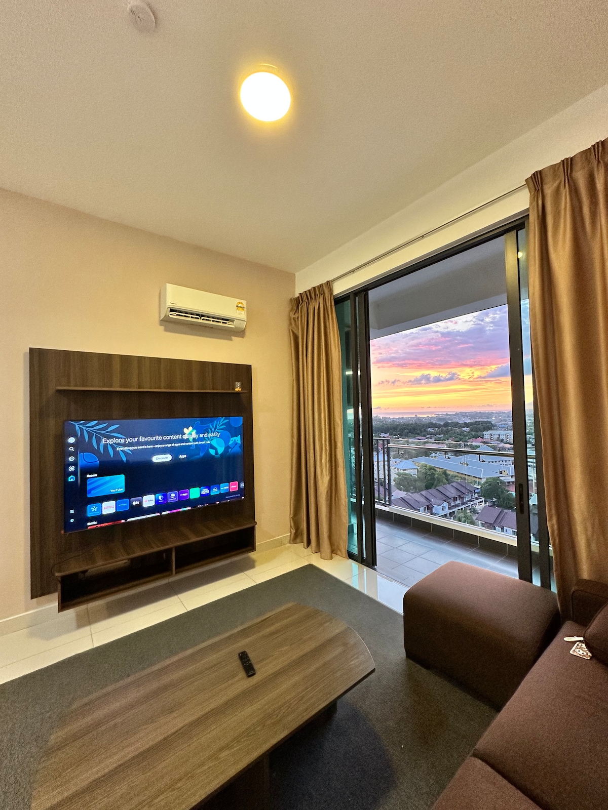 A comfortable living area is showcased, featuring a dark brown sofa and a low wooden coffee table. A flat-screen TV is mounted on the wall, while large glass doors open to reveal a view of a sunset over the city. Light curtains frame the scene.