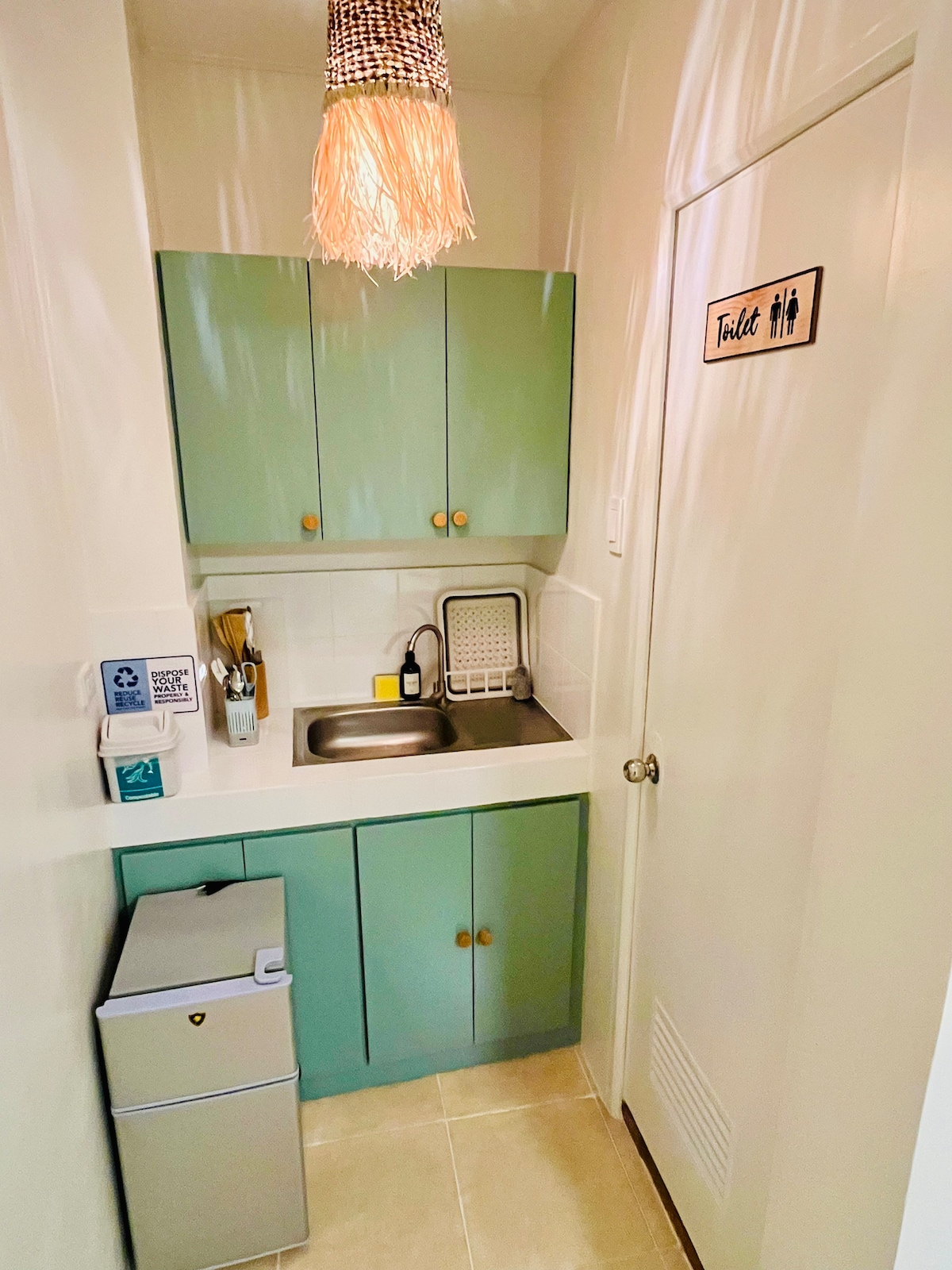 A compact kitchenette is presented with green cabinetry and a stainless steel sink. A small refrigerator is positioned adjacent to the counter, which is complemented by cooking utensils on the countertop. A door labeled 'Toilet' is visible, offering access to additional facilities.