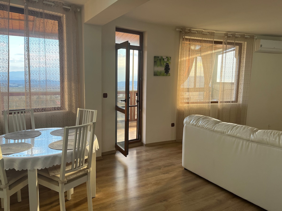 A bright living area features large windows with sheer curtains, providing views of the outdoors. A circular dining table is surrounded by four white chairs, while a cozy white sofa is positioned nearby. A glass door opens to a terrace, enhancing natural light in the space.