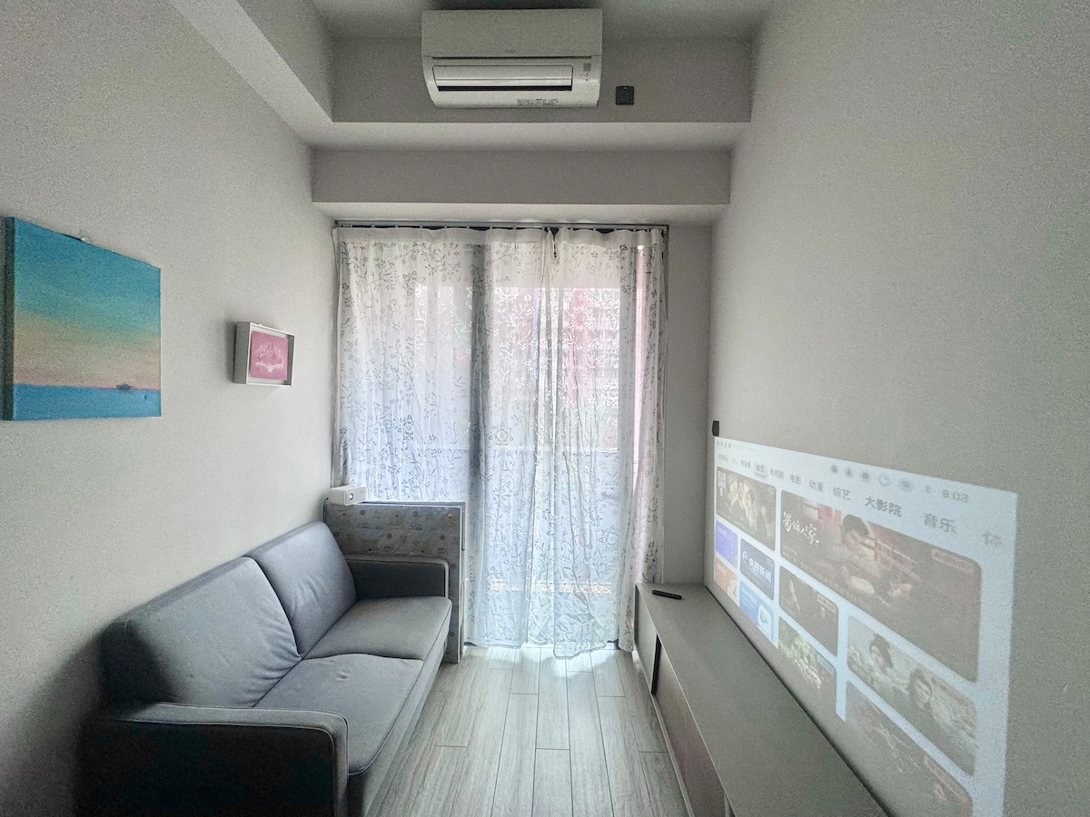A bright and airy sitting area features a gray sofa positioned beside a large window draped with light curtains, allowing natural light to filter in. A projector displays a selection of movies on a wall, enhancing the entertainment experience in the space.