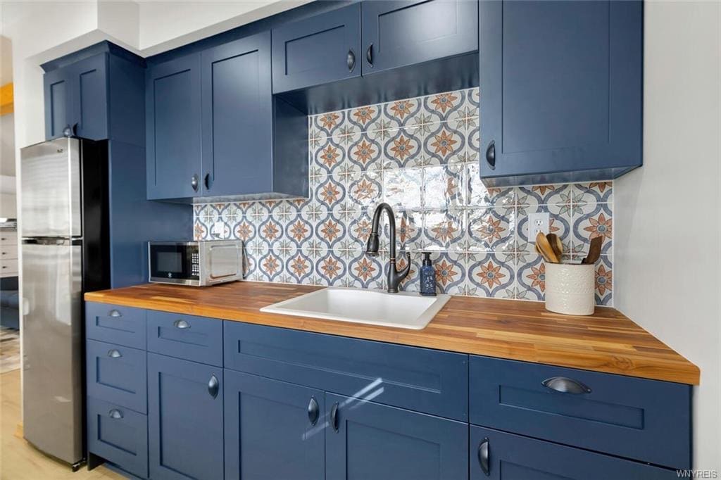 A kitchen area features navy blue cabinetry complemented by a light wood countertop. A large white sink is visible, with modern faucet fixtures. Decorative tiles in soft colors provide a stylish backsplash, while kitchen utensils are organized neatly near the sink.