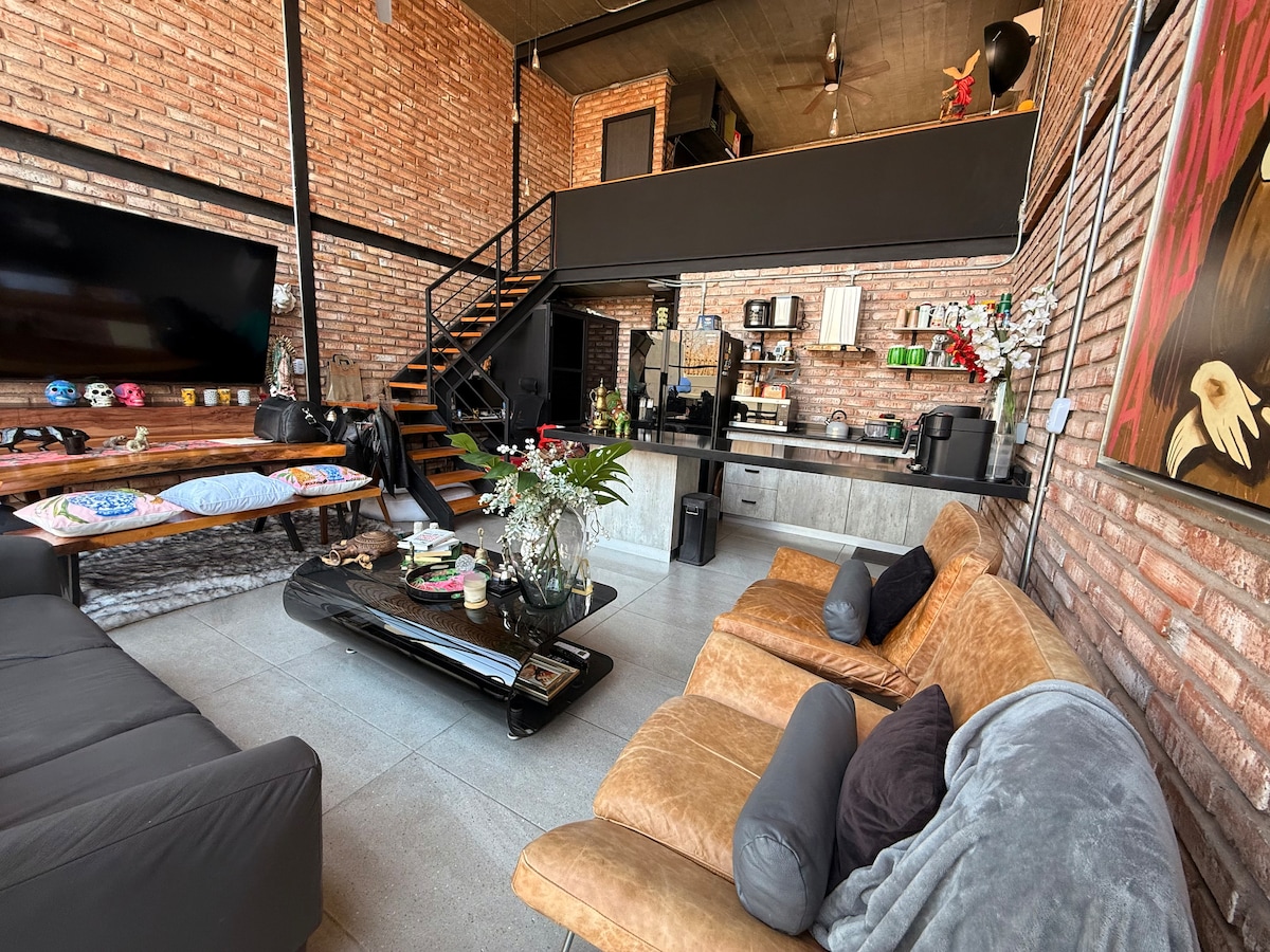 An open living area features exposed brick walls and a high ceiling. A comfortable seating arrangement is complemented by a glass coffee table adorned with a floral centerpiece. A modern kitchen is visible in the background, with a staircase leading to a lofted space.