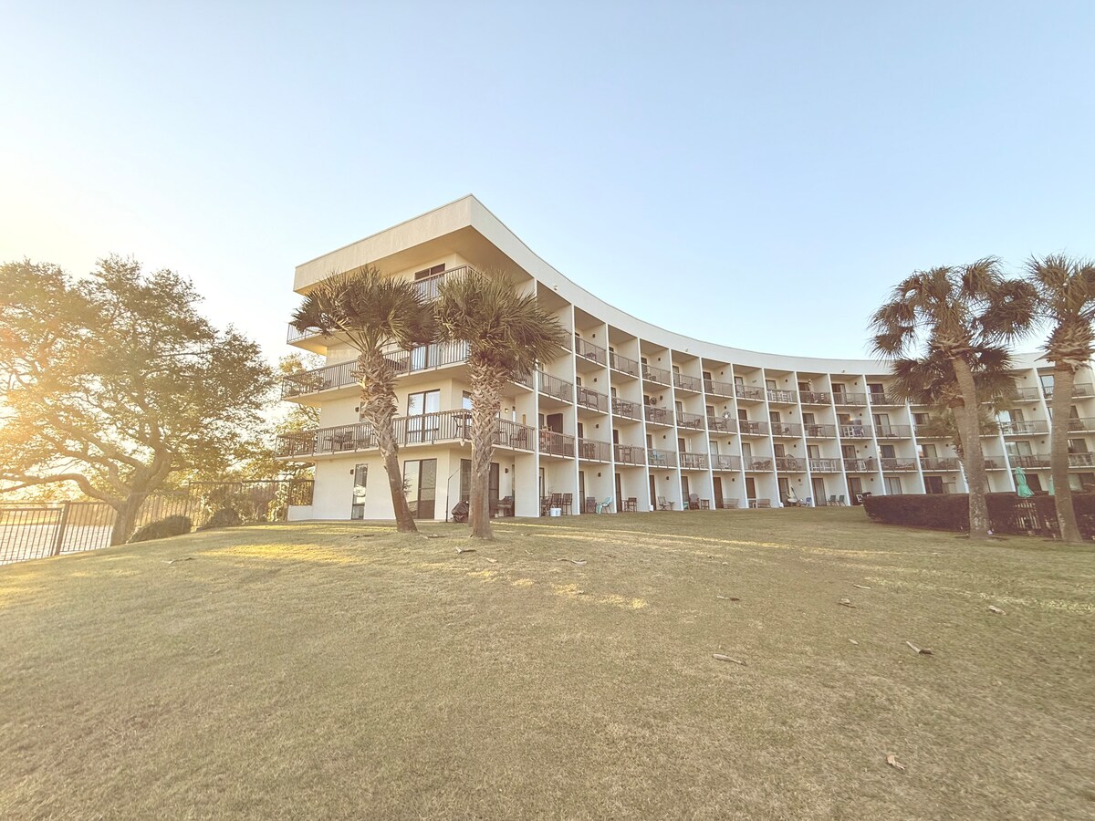 A curved, three-story building is set against a clear sky, featuring multiple balconies with sliding glass doors. Lush greenery, including palm trees, is visible in the foreground, providing a tropical ambiance. The sun casts a gentle glow over the well-kept lawn, enhancing the inviting atmosphere.