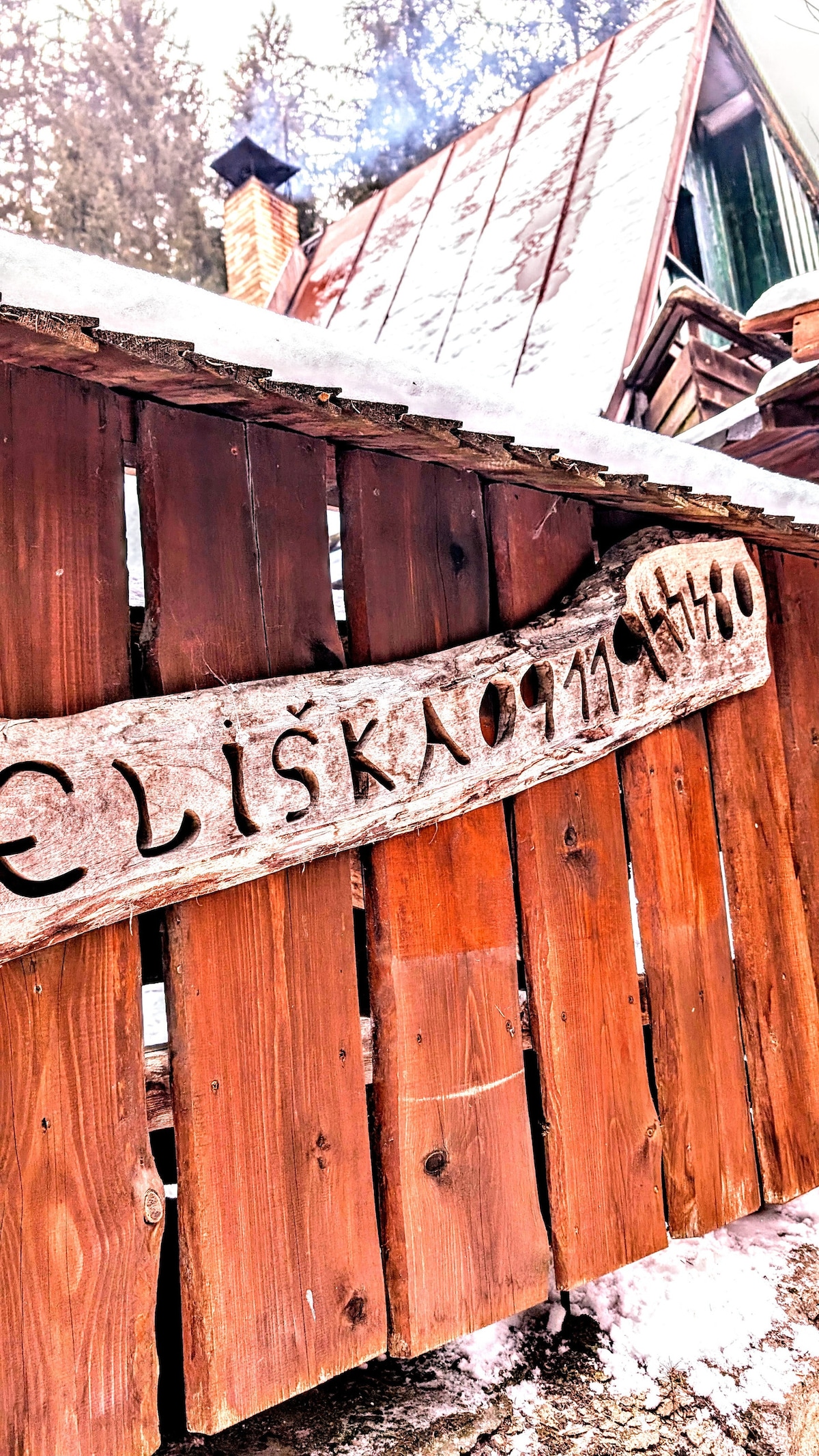 A rustic wooden gate is featured, with a carved sign displaying the name 'Eliška'. The natural texture of the wood contrasts with the metal elements of the gate, surrounded by a snow-covered pathway and trees in the background.
