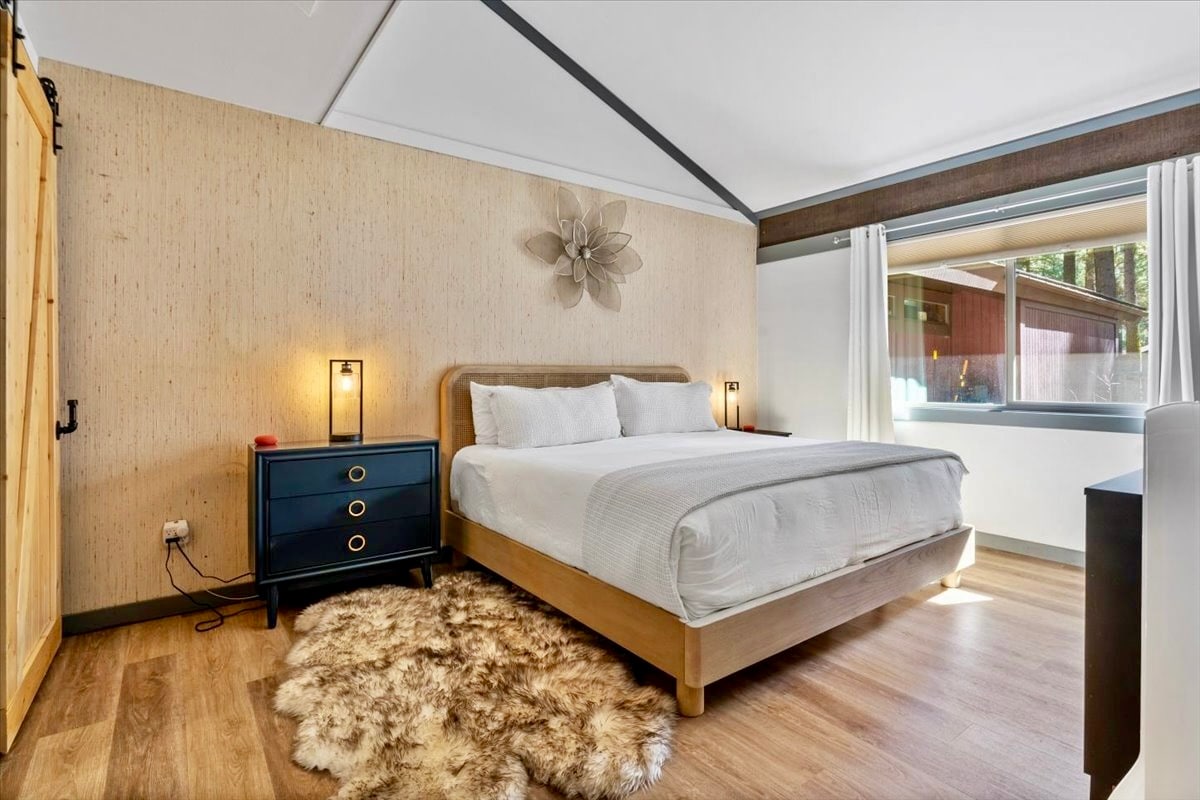 A spacious bedroom features a comfortable bed with crisp linens. Two bedside lamps provide soft lighting, and a dark blue nightstand adds a touch of color. A chair is positioned next to the window, which allows natural light to fill the room.
