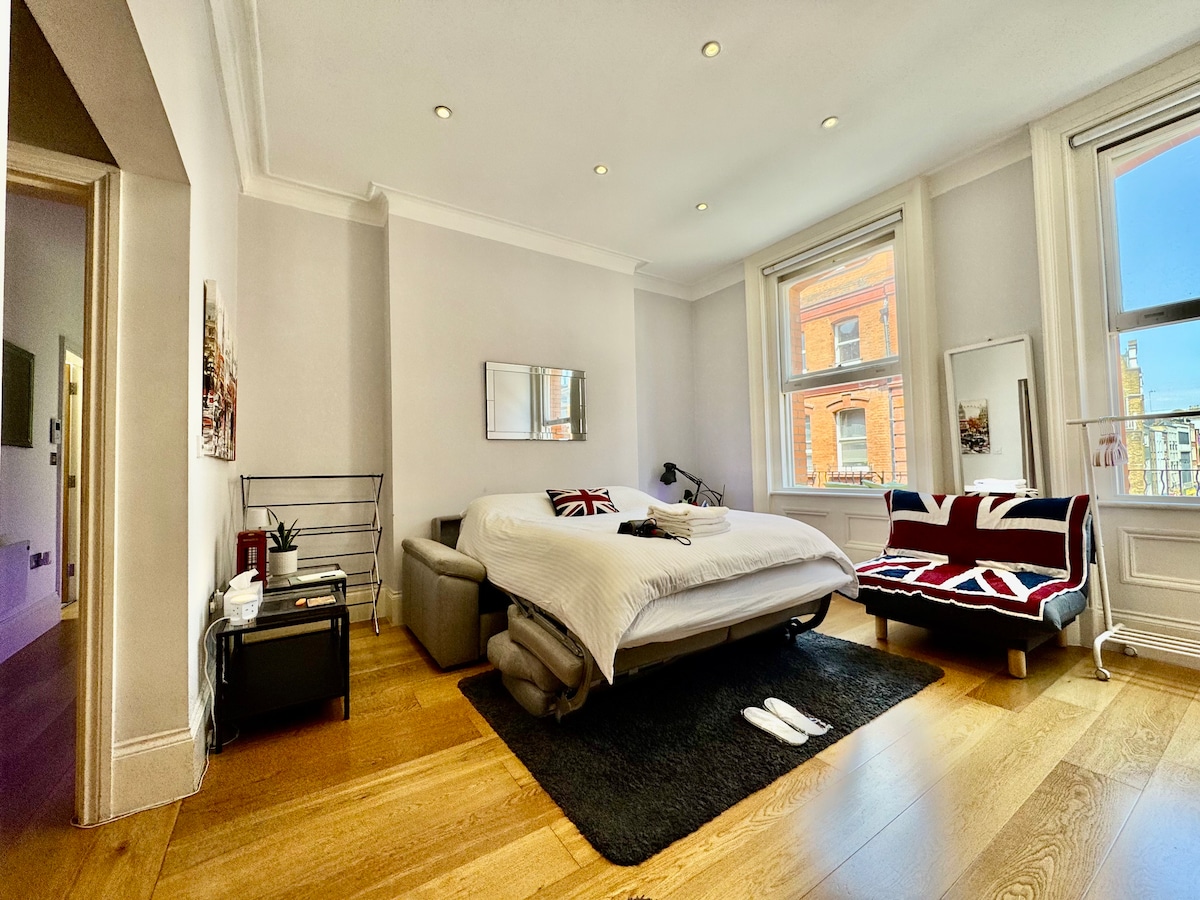 The spacious living area features a queen-sized sofa bed draped in white bedding, alongside a comfortable chair with a Union Jack design. Two large windows allow ample natural light to fill the room, highlighting the wooden flooring and minimalistic decor.