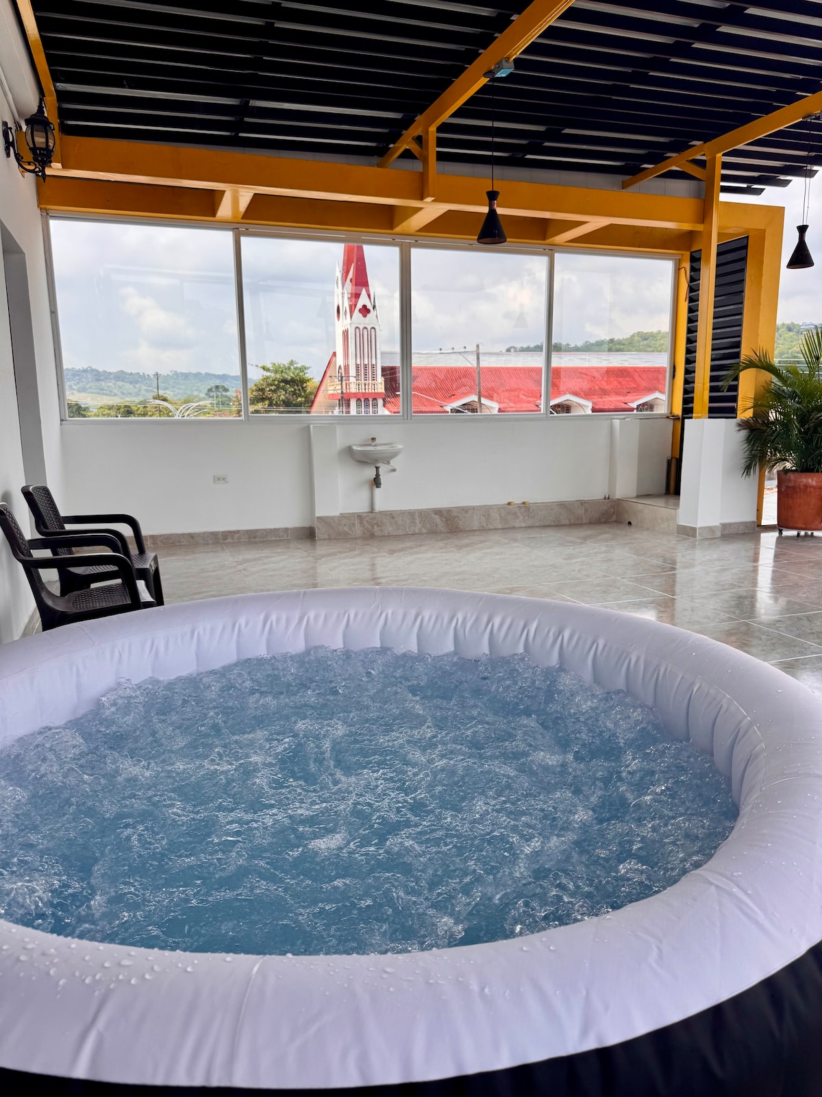 A spacious terrace showcases an inflatable jacuzzi filled with blue water, inviting relaxation. Large windows offer views of the surrounding area, including a red-roofed church. Two black chairs are positioned to enjoy the setting, contributing to a comfortable atmosphere.