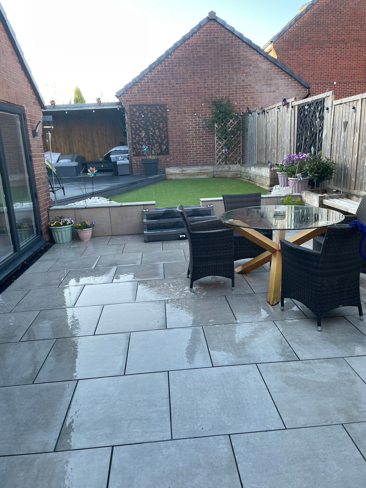 A paved outdoor patio features a wooden dining table surrounded by dark wicker chairs. Potted plants add greenery to the area, while a neatly maintained lawn is visible towards the back. A lounge area with comfortable seating is situated against a wooden backdrop.