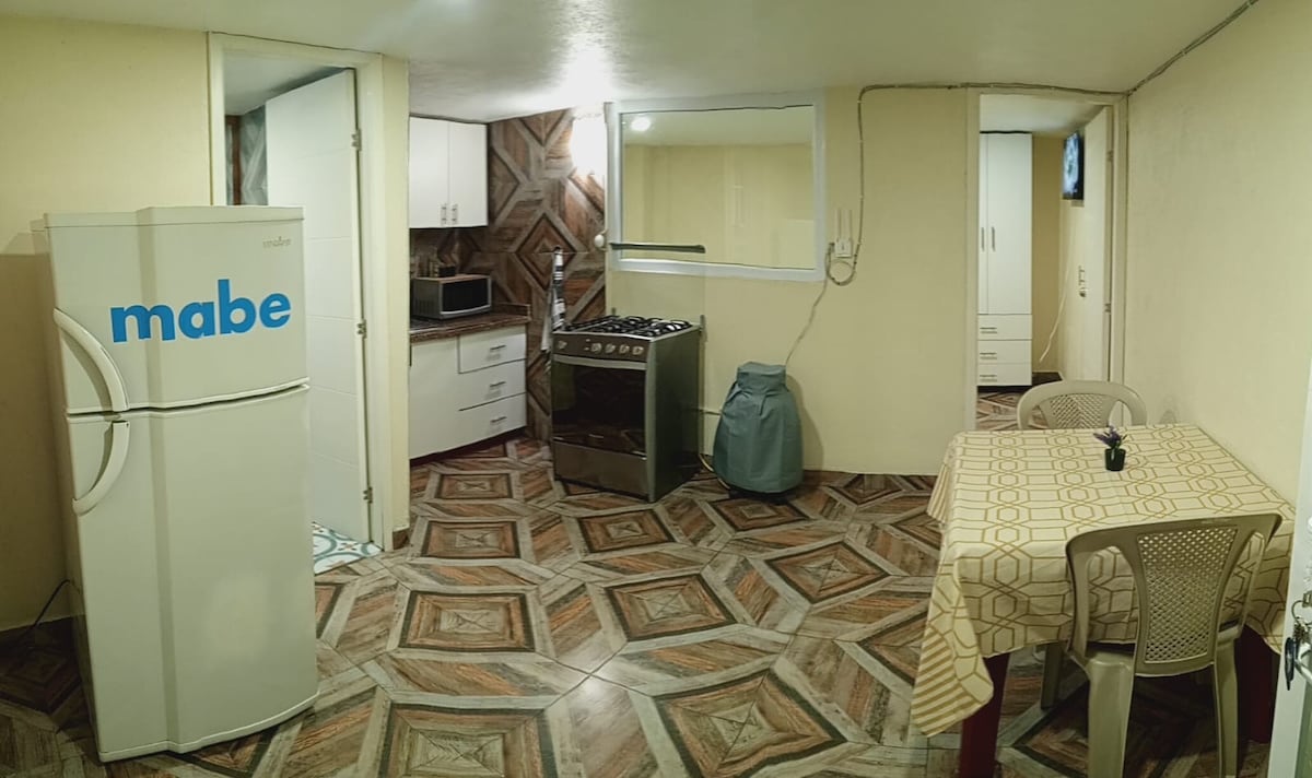 A compact kitchen area is visible, featuring essential appliances including a refrigerator, oven, and microwave. A small table with chairs is placed nearby, while the unique tiled flooring adds texture to the space. Cabinets provide storage and organization.