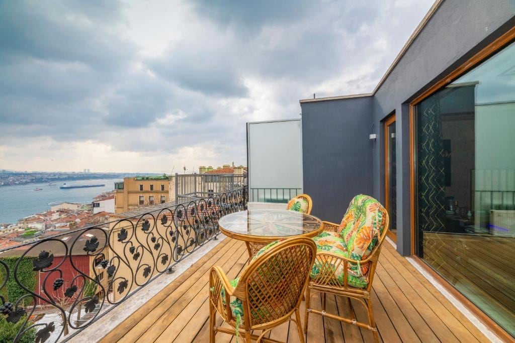A terrace is featured with a glass table and two comfortable chairs, surrounded by decorative railings. A view of the water and cityscape is visible in the background, while the wooden flooring complements the outdoor setting.
