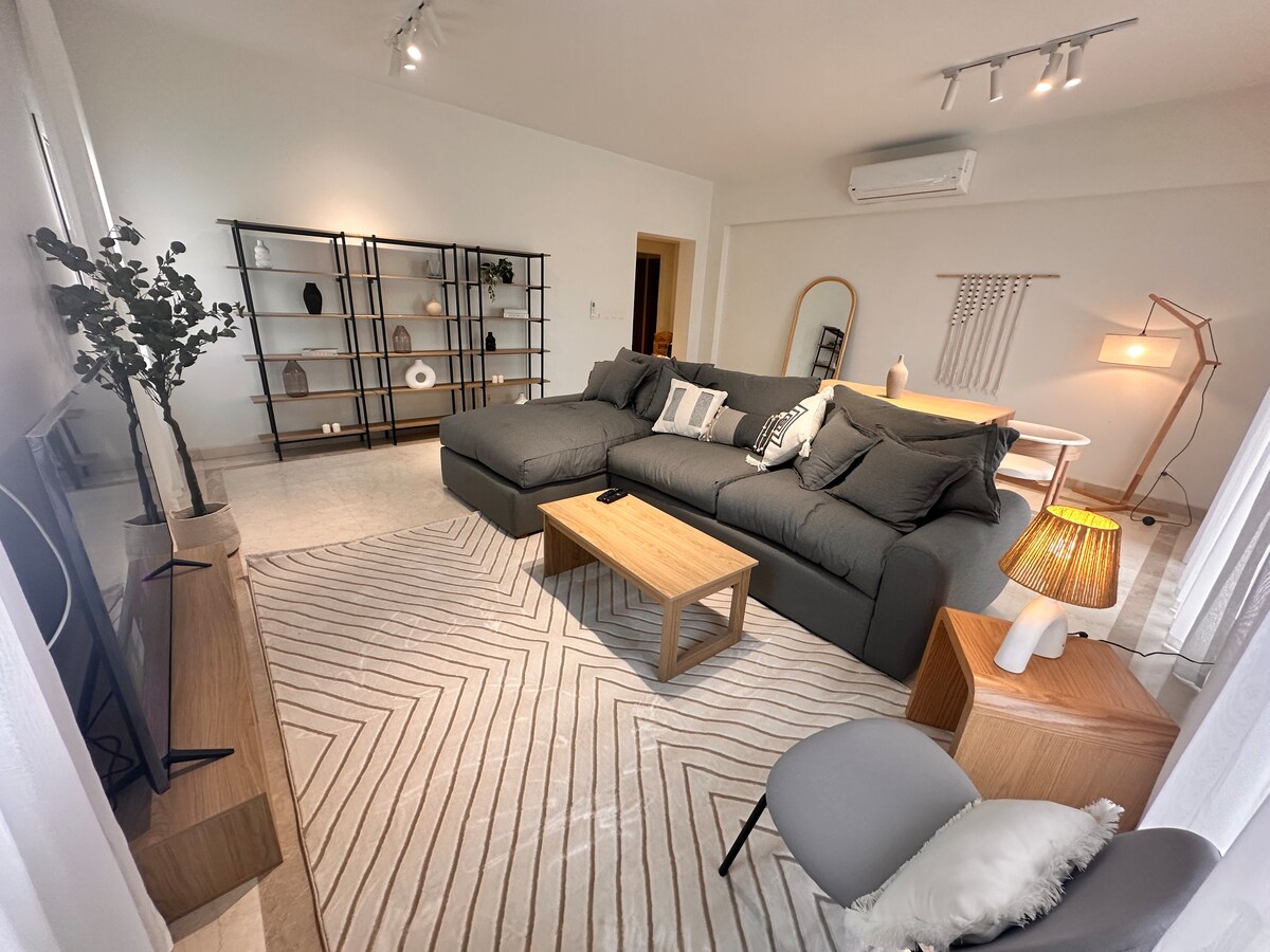 A modern living area is depicted with a spacious sectional sofa arranged around a wooden coffee table. A cozy rug with a geometric pattern covers the floor. In the background, shelves display various decorative items, while soft lighting creates a comfortable ambiance.