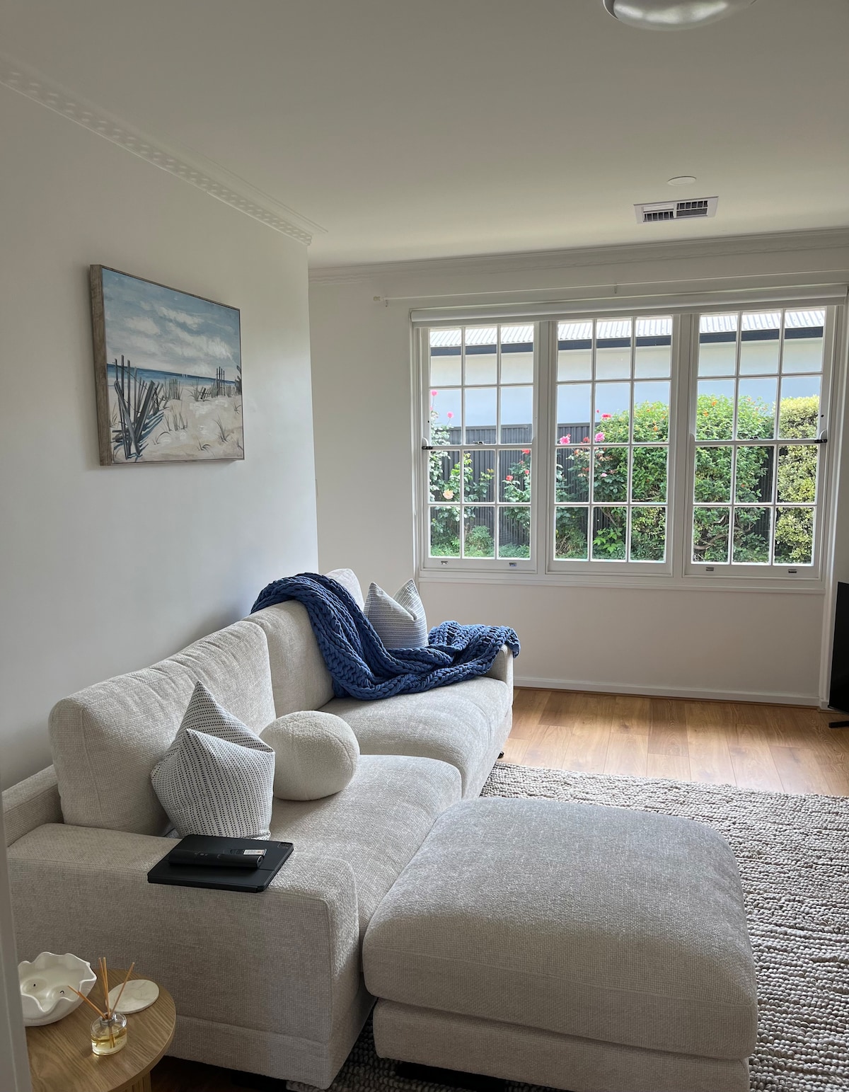 A light-filled living area features a comfortable sectional sofa adorned with neutral-colored cushions and a textured throw. A small coffee table holds decorative items, and a piece of wall art depicting a beach scene adds a serene touch. Large windows invite natural light and offer a view of greenery outside.