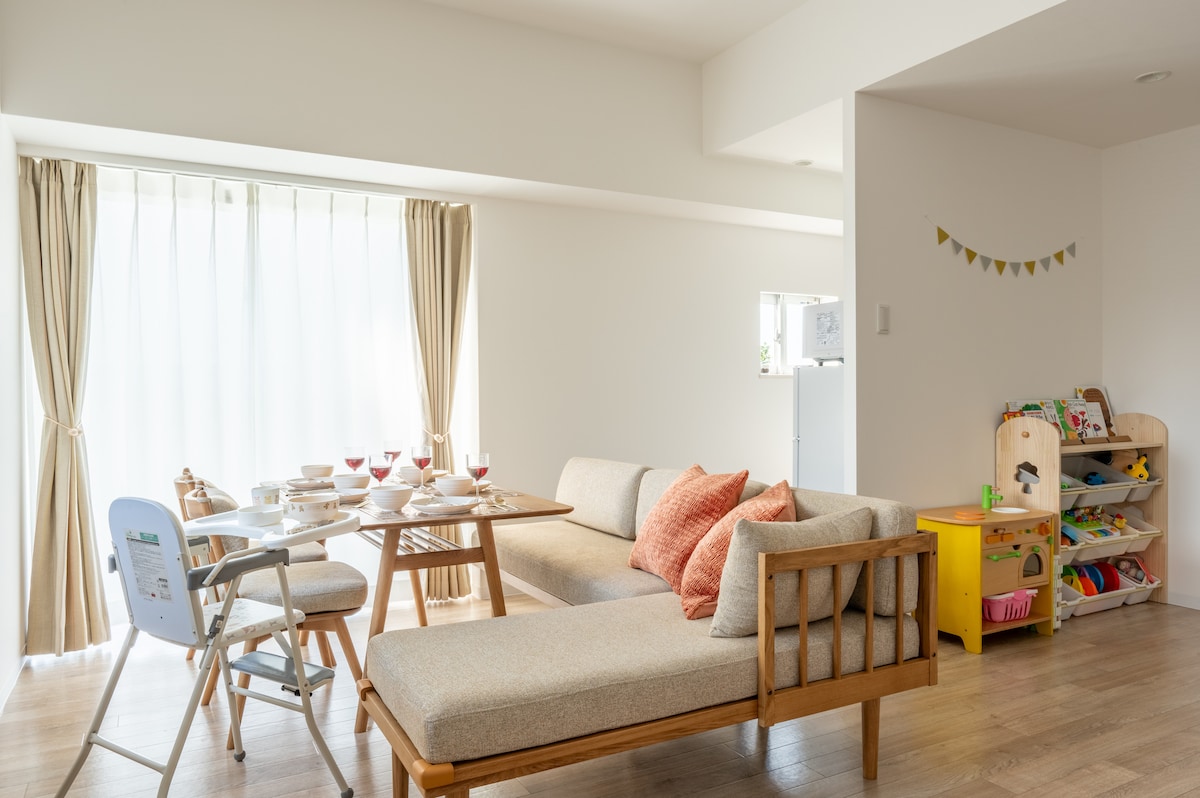 A spacious living area is equipped with a comfortable sofa adorned with cushions. A dining table set for several guests is positioned nearby. In the corner, a designated play area features a colorful assortment of toys, inviting children to enjoy their time.
