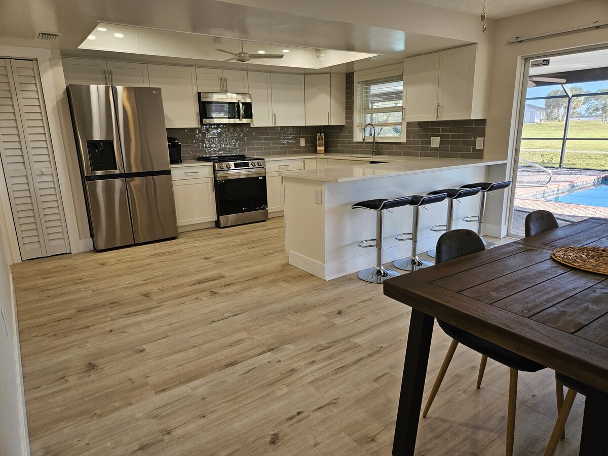 A modern kitchen features stainless steel appliances, including a refrigerator and stove. A large island with seating for four is positioned centrally. Light wood flooring enhances the open layout, which leads to a dining area and offers views of the outdoor space through a sliding glass door.