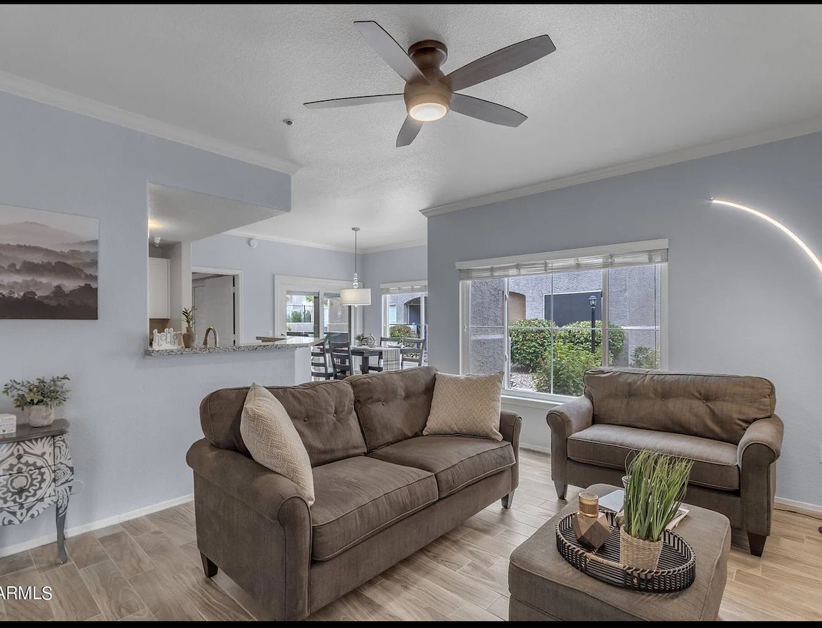 A comfortable living area features two sofas arranged around a round coffee table. A ceiling fan provides ventilation, while natural light floods in through large windows, offering views of the outdoors. The adjacent dining area is partially visible in the background.