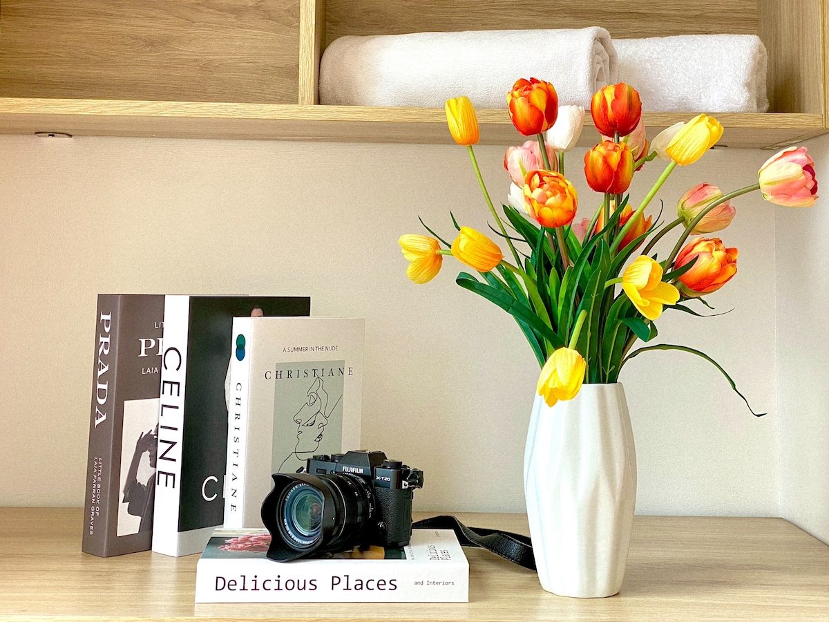 A neatly arranged shelf features a white vase filled with colorful tulips. Nearby, a stack of books titled 'Delicious Places' and various fashion-themed volumes are placed on a wooden surface, along with a camera, creating a cozy, inviting display.
