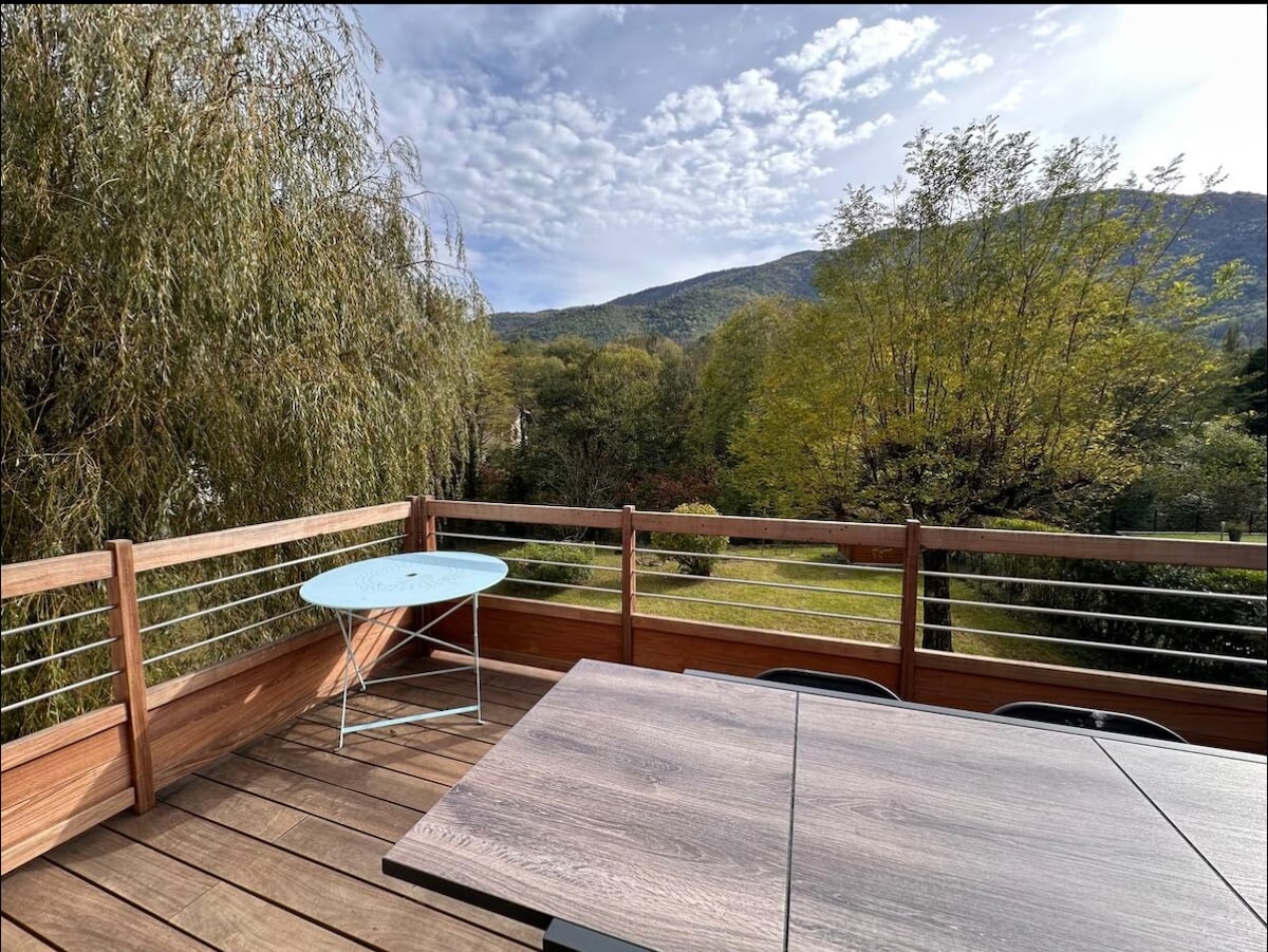 A wooden deck overlooks a serene landscape, featuring green hills and trees under a partly cloudy sky. A light blue table rests on the decking, accompanied by dark chairs, creating a cozy outdoor dining area.