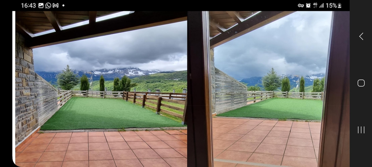 An outdoor terrace is presented, featuring a green lawn area bordered by wooden fencing. The view showcases rolling hills and snow-capped mountains under a cloudy sky. The tiled flooring adds a functional touch to the space, providing a seamless transition from indoors to outdoors.