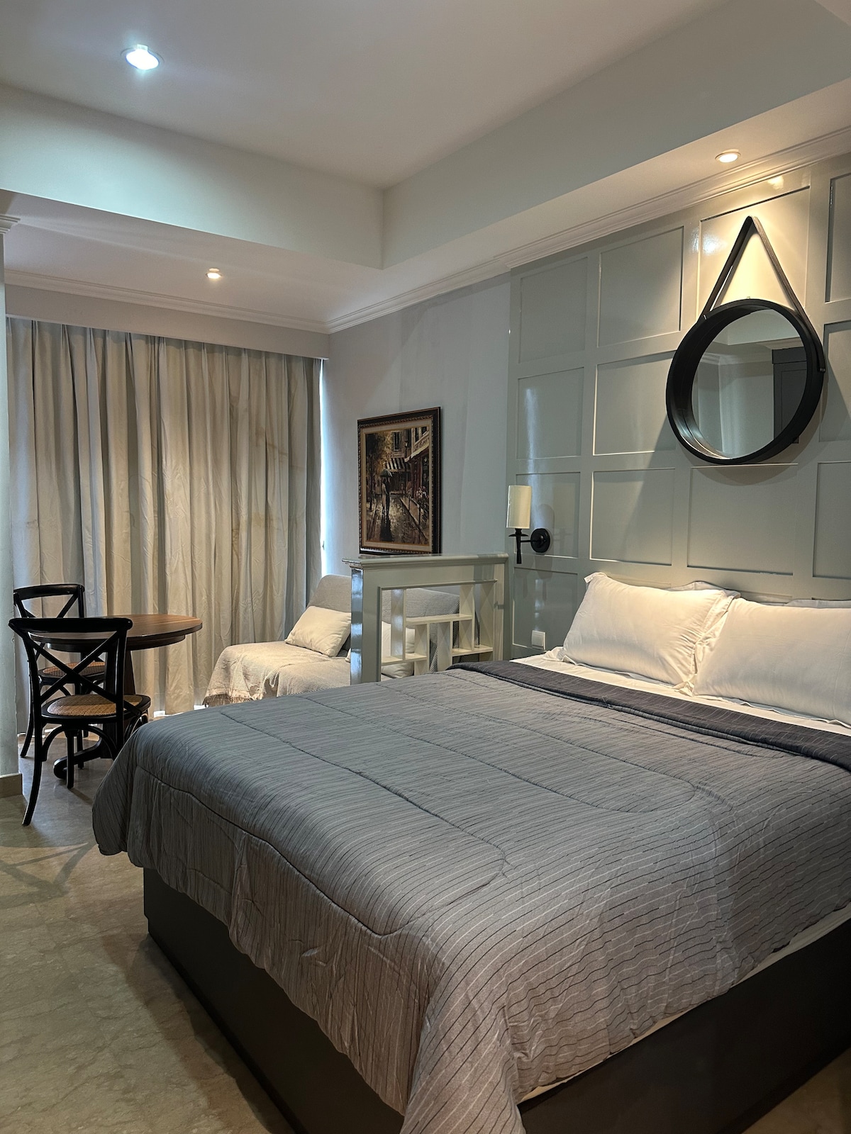 A bright studio apartment features a spacious queen bed with soft linens and a textured throw blanket. A round table with two chairs provides a dining area, while a cozy armchair sits nearby. Natural light filters through sheer curtains, illuminating the modern decor.