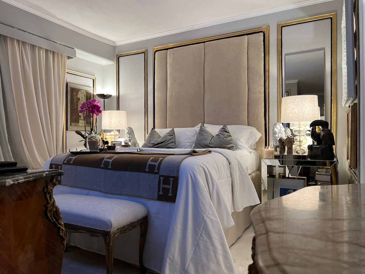 A luxurious sleeping area features a plush bed with an upholstered headboard. Soft lighting from bedside lamps creates a serene ambiance. Decorative pillows complement a stylish throw blanket placed across the bed, while elegant artwork and fresh flowers enhance the sophisticated decor.