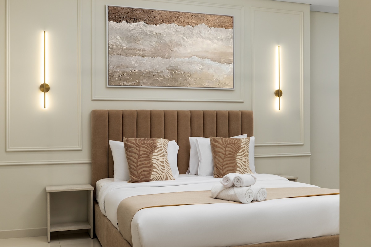 Кадифениот брачен кревет е украсен со стилски перничиња, додавајќи елеганција и удобност. Меките текстури и неутралните тонови создаваат спокојно одморалиште, совршено за опуштање по еден ден во градот.