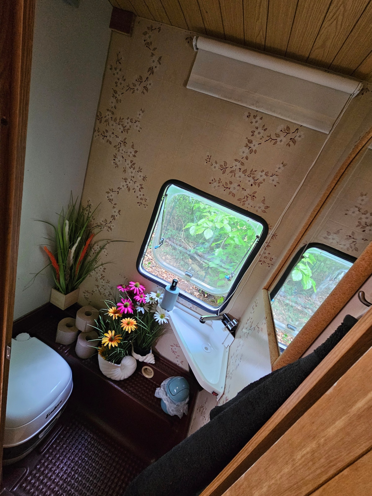 The bathroom features a camping toilet with a flush system, a small sink with a faucet, and a window offering views of the greenery outside. Decorative plants and flowers add a touch of color, while essential toiletries and towels are neatly arranged.