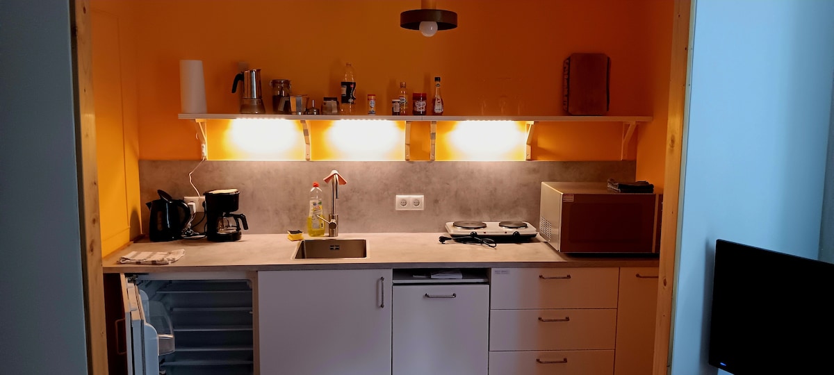 The kitchen features a blend of modern appliances and ample storage. A refrigerator is positioned to the left, with a microwave and a double hot plate on the counter. Under-cabinet lighting enhances visibility over the workspace, which includes a sink and various kitchen essentials.