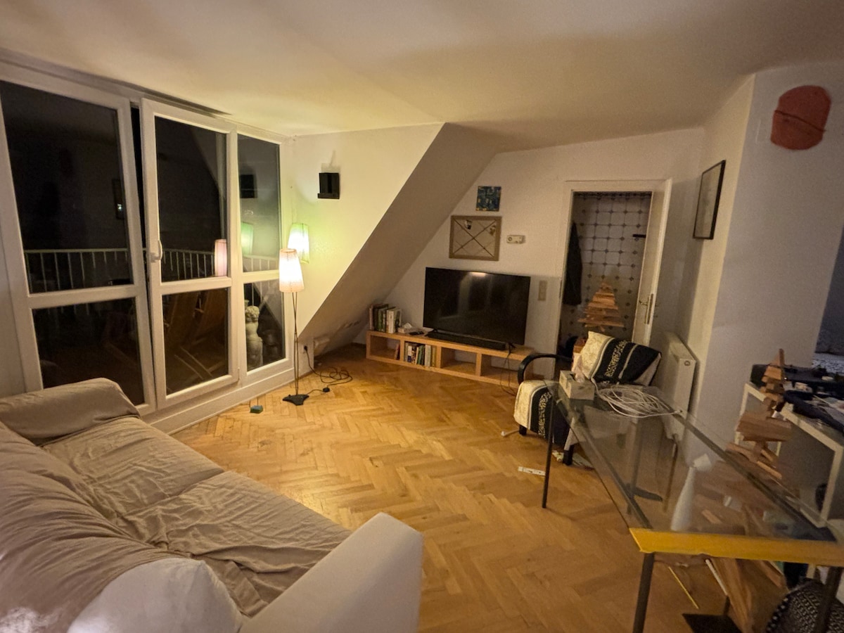 A spacious living area features large glass windows allowing natural light to fill the room. A flat-screen TV is placed on a wooden console, while a comfortable sofa and chairs are arranged invitingly. A lamp with a soft glow adds warmth to the space.